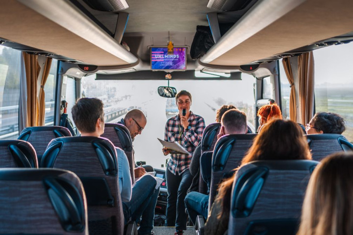 Skupina ljudi se pelje na avtobusu, vodička govori po mikrofonu, ostali jo poslušajo ali gledajo skozi okno.