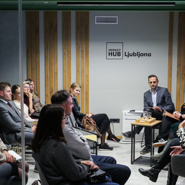 Obiskovalci ob poslušanju štirih govorcev na odru, v ozadju projekcija